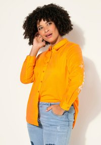 Camicia arancione a maniche lunghe con bottoni e colletto, con accenti grafici bianchi sulle maniche. Indossata con jeans chiari strappati.