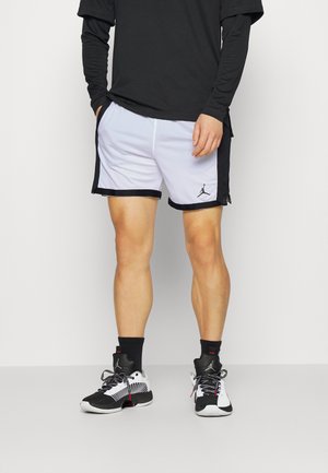 Hombre vistiendo pantalones cortos de baloncesto blancos y negros, camiseta negra de manga larga, calcetines negros y zapatillas de baloncesto en blanco y negro, de pie sobre un suelo gris.