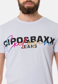 Weißes Baumwoll-T-Shirt mit schwarzer, roter, blauer und goldener Stickerei, die "GIPOSBAXX Exclusive Jeans" zeigt. Kurze Ärmel, Rundhalsausschnitt.