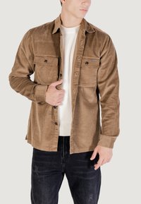 Giacca camicia in corduroy marrone con due tasche sul petto, chiusura con bottoni e texture a coste, indossata sopra un maglione bianco a maglia e jeans neri.