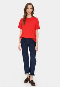 Tricou roșu cu mâneci scurte asociat cu blugi din denim închis, răsuciți la gleznă, și pantofi mocasi maro cu detalii din franje. O ținută simplă, casual.