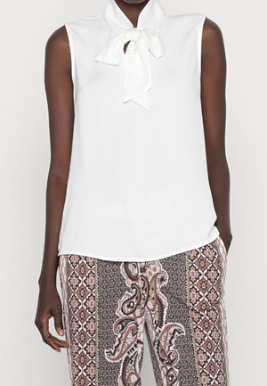 Blouse blanche sans manches avec un col noué en nœud papillon, associée à un pantalon taille haute avec un motif cachemire marron et noir et floral.