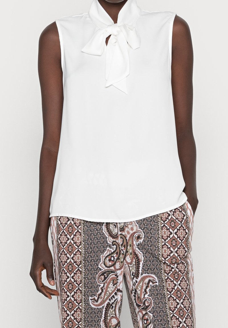 Blouse blanche sans manches avec un col noué en nœud papillon, associée à un pantalon taille haute avec un motif cachemire marron et noir et floral.