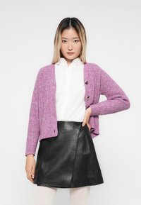 Ung kvinne med rett blondt hår iført en hvit knappeskjorte, lilla cardigan, svart skinnskjørt, og poserer med en hånd på hoften.