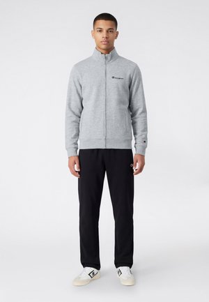 Giovane uomo che indossa una felpa grigia con zip, pantaloni neri e sneakers bianche con loghi di marca, in piedi contro uno sfondo chiaro e semplice.