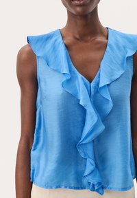 Blouse sans manches bleu clair avec un décolleté en V à volants. Fabriquée dans un tissu lisse avec une texture douce et une coupe ample.