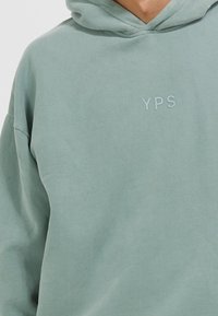 Sweat à capuche vert clair avec "YPS" imprimé en petites lettres sur la partie supérieure de la poitrine, porté par une personne avec le cou et les épaules partiellement visibles.