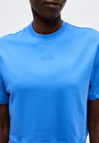 Κοντινό ενός ατόμου που φοράει φωτεινό μπλε μπλουζάκι με μικρό ανάγλυφο λογότυπο "BORG" στο στήθος, περικομμένο κάτω από τη μύτη.