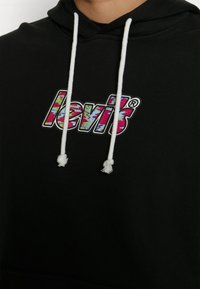 Sweat à capuche noir avec cordons blancs et logo Levi's coloré imprimé sur la poitrine.