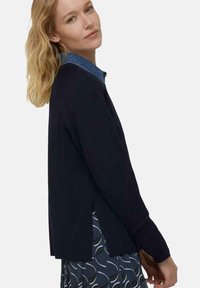 Marineblaues Sweater mit seitlichen Schlitzen, langen Ärmeln und einem Denim-Kragen; getragen über einem gemusterten Rock mit wellenförmigen Designs in dunklen Farben.