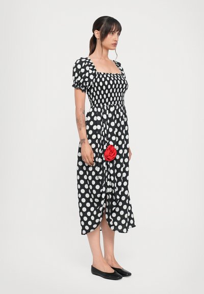 Rochie alb-negru cu buline, cu mâneci bufante, un corset ajustat și un detaliu din floare roșie la tiv. Purtați cu pantofi joși negri.