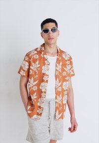Camisa de manga corta, de color naranja con estampado floral de hibiscos blancos, llevada sobre una camiseta sin mangas blanca, combinada con pantalones cortos gris claro.