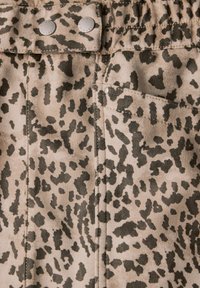 Beige mokka-kangas, jossa on musta pilkullinen kuvio, kaksi metallista nappia ja sivutasku. Pehmeä rakenne, jossa on näkyvät saumat.