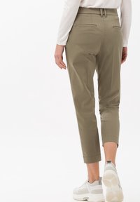 Kobieta w beżowych spodniaczkach i białych sneakersach, stojąca plecami do aparatu na jednolitym białym tle.