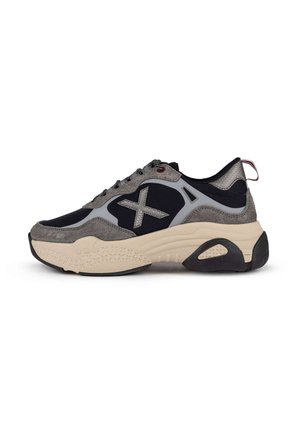 Zapatilla chunky con parte superior de malla negra, acentos en gamuza gris y caucho, mediasuela crema y suela exterior de caucho negro texturizado. Lengüeta roja en el talón.