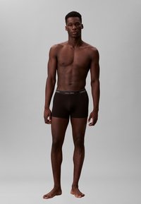 Männlicher Model steht barfuß vor grauem Hintergrund, trägt schwarze Calvin Klein Boxershorts und blickt mit neutralem Ausdruck geradeaus.