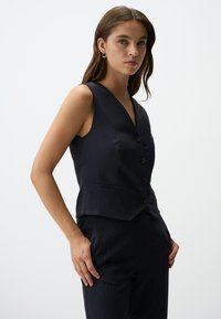 Gilet a righe navy con scollo a V, cinque bottoni marroni e due tasche frontali, realizzato in un tessuto strutturato. Design minimalista.