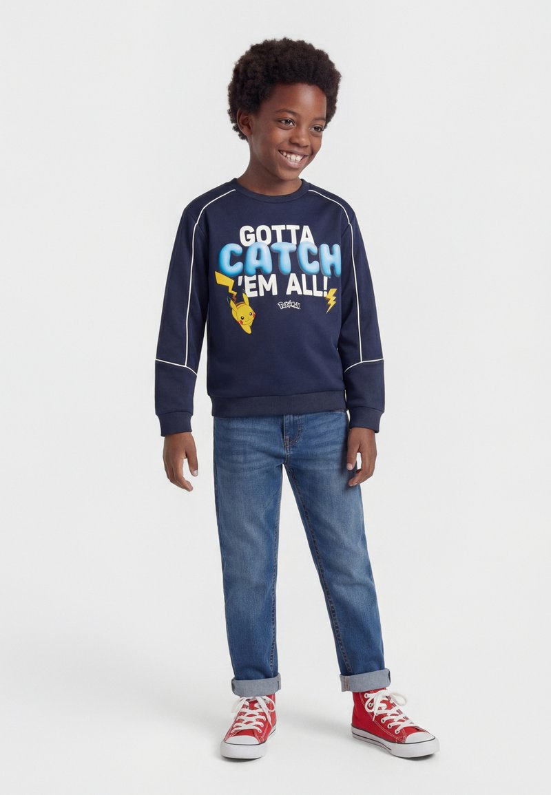 Poiss kannab tumesinist dressipluusiga, millel on kiri "Gotta Catch 'Em All" ja Pikachu pilt, siniseid teksapükse ning punaseid kõrge säärega vabaajajalatsid, naeratades ja seistes.