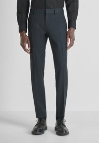 Man in slim-fit donkergrijze broek, zwarte dresschoenen en een ingestopte zwarte blouse met lange mouwen tegen een effen achtergrond.