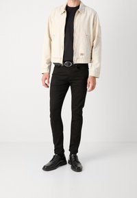 Bežová džínová bunda s límcem, předními kapsami a světlým prošitím, kombinovaná s černým tričkem a úzkými černými džíny, doplněná černými botami.