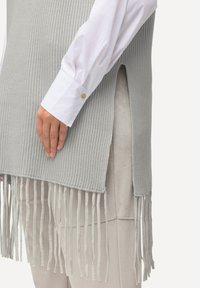 Světle šedý ribbed pletený svetr s bočními rozparky a střapci na spodním lemu, vrstvený přes bílou košili s knoflíky. Hladká textura, ležérní design.