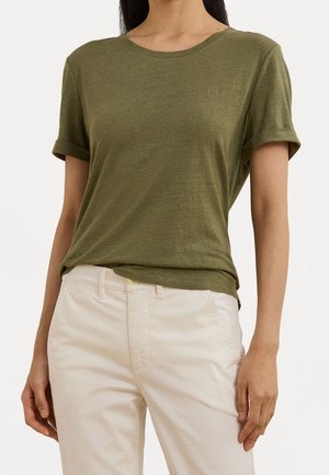 T-shirt vert olive à manches retroussées, col rond et détail discret du logo. Associé à un pantalon de couleur claire. Texture douce.