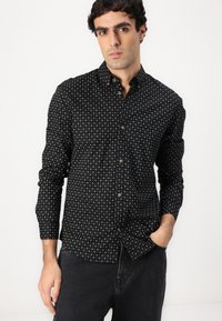 Schwarzes Hemd mit Knöpfen und kleinem weißen Kreuzmuster, lange Ärmel und Kragen. Der Stoff wirkt glatt und leicht. Kombiniert mit dunklen Jeans.