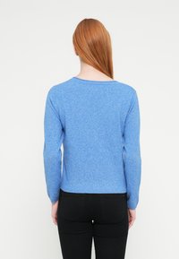 Zils adīts džemperis ar garām piedurknēm, apaļu kakla izgriezumu un vieglu tekstūru, kombinēts ar melnām cieši piegulošām biksēm, skatīts no aizmugures.