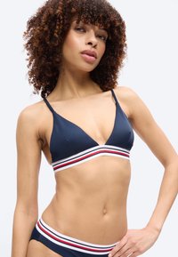 Γυναίκα με ξανθά σγουρά μαλλιά που φοράει μπικίνι σε ναυτικό μπλε με λευκές, κόκκινες και μαύρες ριγέ ζώνες, ποζάροντας μπροστά σε ένα απλό φωτεινό φόντο.
