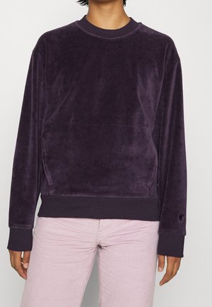 Persoon met een donkerpaarse velours trui met lange mouwen en lichtroze ribfluwelen broek, staand tegen een effen achtergrond.