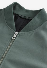 Gros plan sur le col d'une veste verte avec une fermeture éclair argentée et une doublure intérieure noire à motif texturé.