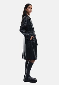 Czarny skórzany płaszcz trench z paskiem w talii, długimi rękawami, bocznymi kieszeniami i płynącym krojem, w połączeniu z czarnymi botkami do kolan.