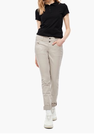 Kobieta ubrana w czarną koszulkę polo z krótkim rękawem, beżowe spodnie z podwiniętymi nogawkami, białe skarpetki i białe trampki, stoi z jedną ręką w kieszeni.