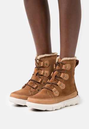 Botas de tornozelo em camurça castanha com forro em pelo sintético, seis ilhós, atacadores castanhos e sola de borracha branca. Projetadas para conforto e estilo.