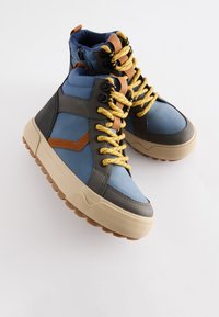 Sneaker alte con un design blu e grigio, materiali texturizzati, lacci gialli, dettagli marroni e una robusta suola in gomma.