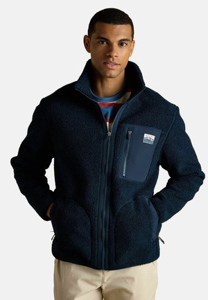 Mann trägt eine marineblaue Sherpa-Fleecejacke mit Reißverschlusstasche auf der Brust, gestreiftes Hemd darunter, beige Hose, Hände in den Seitentaschen, neutraler Hintergrund.