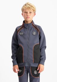 Blond kind gekleed in een marineblauw en zwarte trainingsoutfit met oranje bies, die recht vooruit kijkt tegen een witte achtergrond.