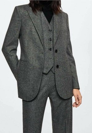 Femme portant un costume gris trois pièces avec une veste, un gilet boutonné et un pantalon par-dessus un col roulé noir.