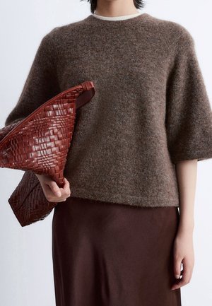 Person iført en fluffy brun kortærmet sweater og brun nederdel, der holder en stor vævet mørkerød læder clutch i hånden.
