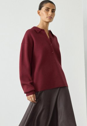 Femme portant un pull long à boutons bordeaux et une jupe en cuir marron foncé, se tenant devant un fond blanc uni.