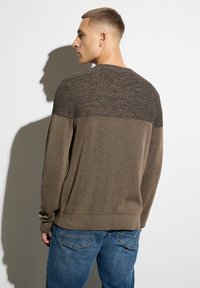 Brauner Pullover mit strukturiertem Oberteil, geripptem Körper und langen Ärmeln. Getragen mit blauen Jeans, die eine entspannte Passform und ein lässiges Design zeigen.