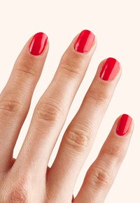 Esmalte de uñas rojo aplicado en cinco dedos, mostrando un acabado brillante en uñas cortas y en forma ovalada sobre un fondo beige claro.