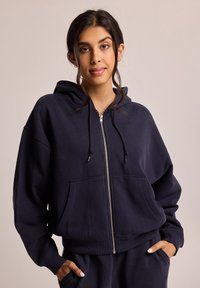 Navy blue zip-up hoodie in soft cotton blend, featuring a front pocket, drawstring hood, and ribbed cuffs. Simple, relaxed fit design.