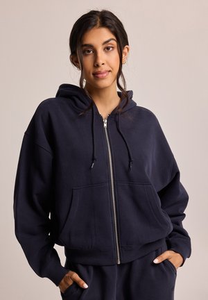 Sudadera con cremallera en azul marino, fabricada en una suave mezcla de algodón, con un bolsillo frontal, capucha con cordón y puños acanalados. Diseño simple y corte relajado.
