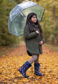 Groene fleecejack, doorzichtige paraplu met zwarte accenten, bruine leggings, blauwe regenlaarzen met een roze hart en blauw patroon, wandelend op gele bladeren.