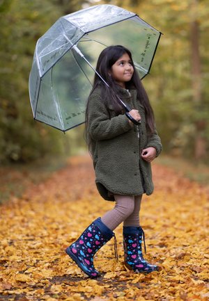 Veste en polaire verte, parapluie transparent avec des accents noirs, leggings marron, bottes de pluie bleues avec un cœur rose et un motif bleu, marchant sur des feuilles jaunes.