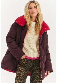 Bordowy pikowany płaszcz z czerwonym kołnierzem, przeszywany wzór, z przodu guziki, zestawiony z kremowym swetrem i wzorzystymi spodniami w panterkę.