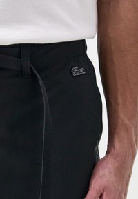 Pantalons noirs en tissu lisse avec un léger éclat. Dotés d'un cordon de taille sur le côté et d'un petit logo brodé sur la poche.