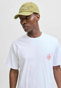 Hvid bomulds T-shirt med et lille rødt broderet design på venstre bryst, parret med en lysegrøn kasket med "Originals" i kursiv.