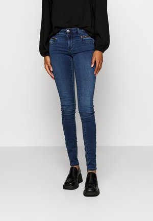 Jeans de mezclilla ajustados de color azul oscuro con una textura suave, que cuentan con bolsillos frontales, una cremallera y un cierre de botón. Combinados con mocasines negros.